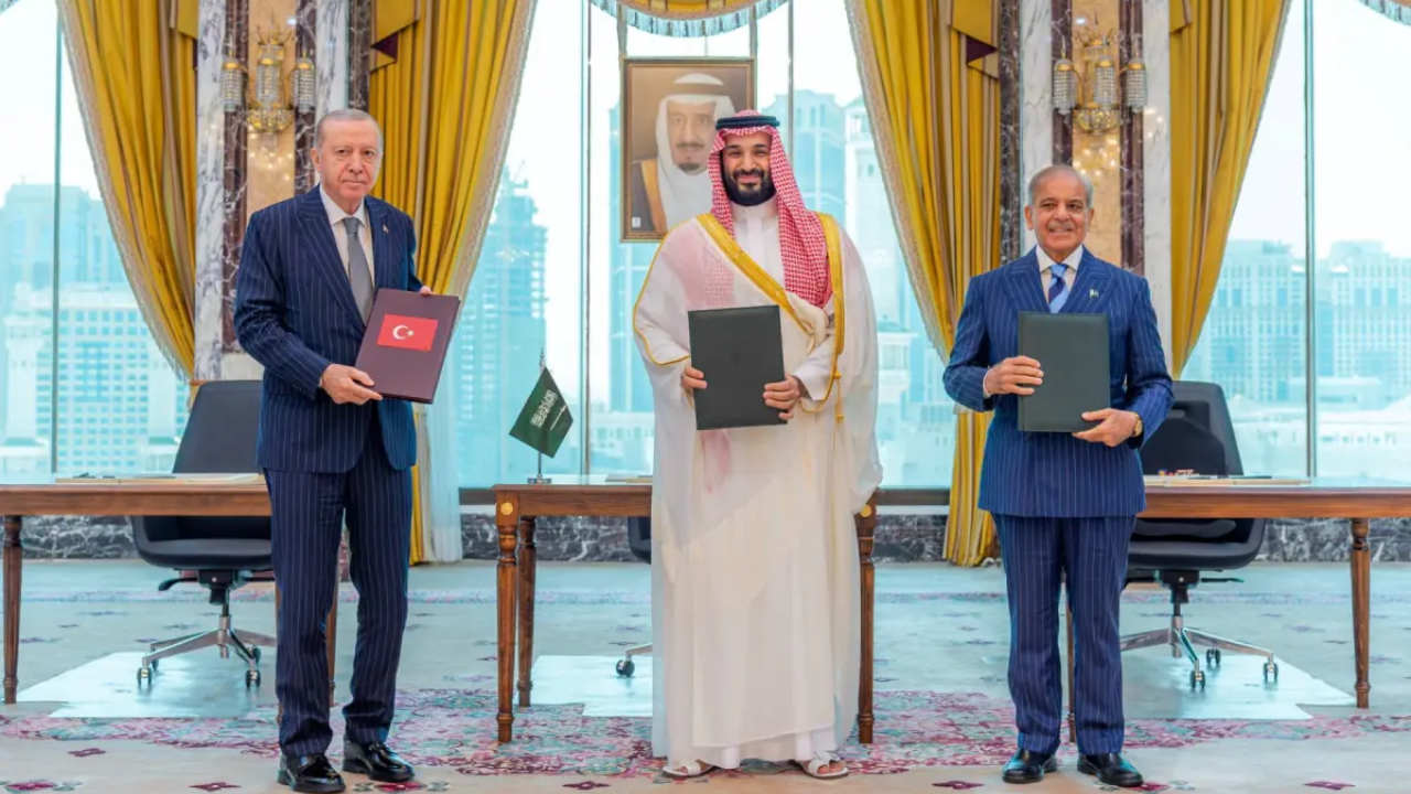 Three leaders standing side by side in a formal ornate office holding signed document folders: Turkish President Recep Tayyip Erdogan on the left holding a folder with the Turkish flag, Saudi Crown Prince Mohammed bin Salman in the center in white thobe and red checkered ghutra holding a black folder, and Pakistan Prime Minister Shehbaz Sharif on the right in a blue pinstripe suit holding a black folder. Behind them are gold curtains, a small Saudi flag on a table, and a framed portrait of King Salman.
