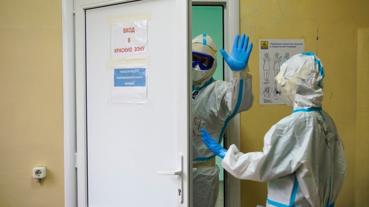 Two medical workers wearing full protective suits and masks stand at the entrance to a restricted hospital area marked with warning signs.