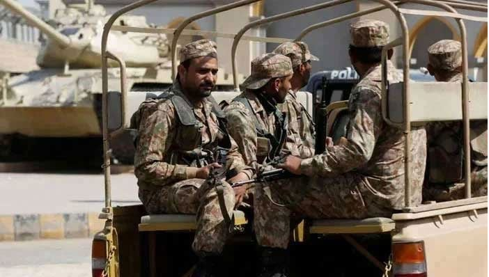 Pakistani soldiers riding in the back of a military vehicle, with a tank visible in the background.