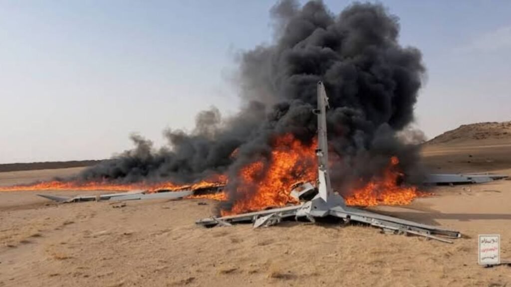 A wrecked aircraft lies on a desert landscape engulfed in flames, with thick black smoke rising into the sky.