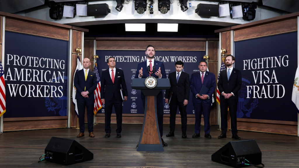 Vice President JD Vance speaking at a podium with the White House seal in a press briefing room, flanked by five men in suits standing behind him. Large dark blue banners on both sides read "PROTECTING AMERICAN WORKERS" and "FIGHTING VISA FRAUD" with smaller text "AMERICAN JOBS" and White House seals in the background. American flags are visible on stage with overhead studio lighting.