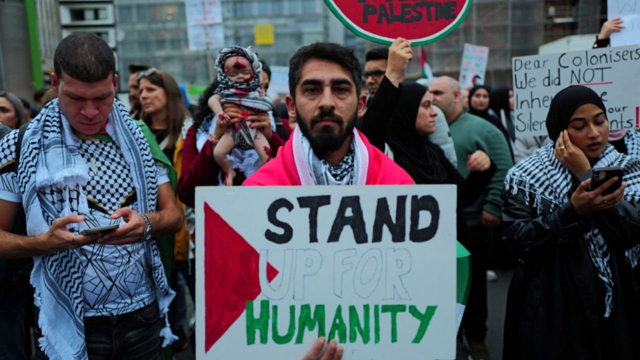 Protesters wearing keffiyehs gather at a demonstration, with a man holding a sign reading “Stand Up for Humanity” and others carrying pro-Palestinian signs and flags.