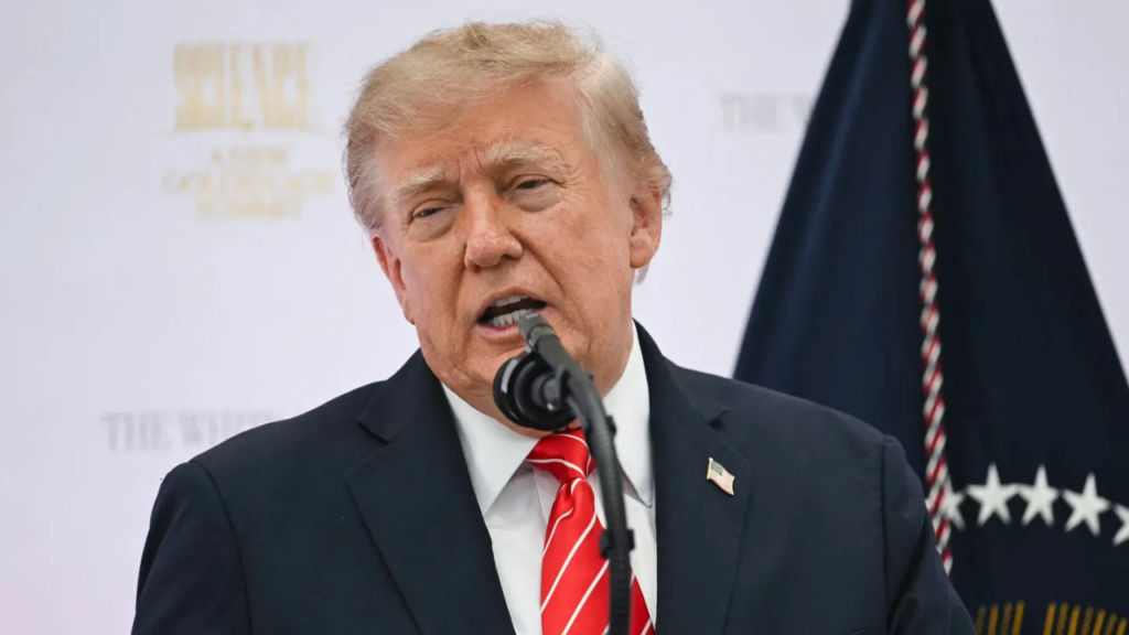 Close-up of U.S. President Donald Trump speaking into a microphone at a podium, wearing a dark navy suit, white shirt, and red tie with white diagonal stripes, with a small American flag pin on his lapel. Behind him is a white backdrop with faint gold text and a dark blue flag with white stars and red-and-white twisted trim visible on the right.