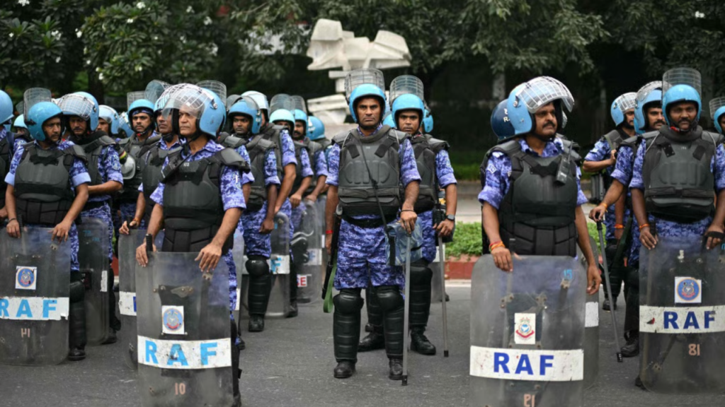 A line of Indian Rapid Action Force (RAF) police officers standing in formation on a street outdoors. They wear blue camouflage uniforms, black body armor, black knee pads, and light blue helmets with clear face shields. Most hold large transparent riot shields with "RAF" written in blue letters and an emblem above it, and some hold batons. Trees and a white abstract sculpture are visible in the background.